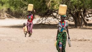 Pictured are two women balancing plastic water jugs on their heads as they walk, one holds the hand of a child who walks alongside her. | Photo credit: Julien Ayroles/ UC Berkeley